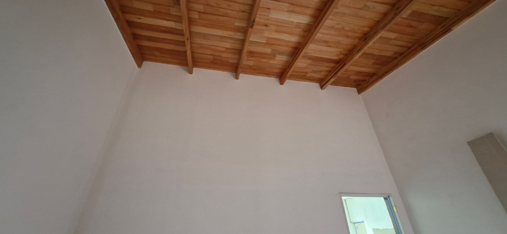 Vista interior de una habitación con techo de listones de madera y paredes blancas. Se ve una pequeña ventana.
