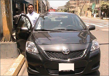 Hombre de pie junto a un sedán Toyota negro descapotable en una calle de la ciudad.