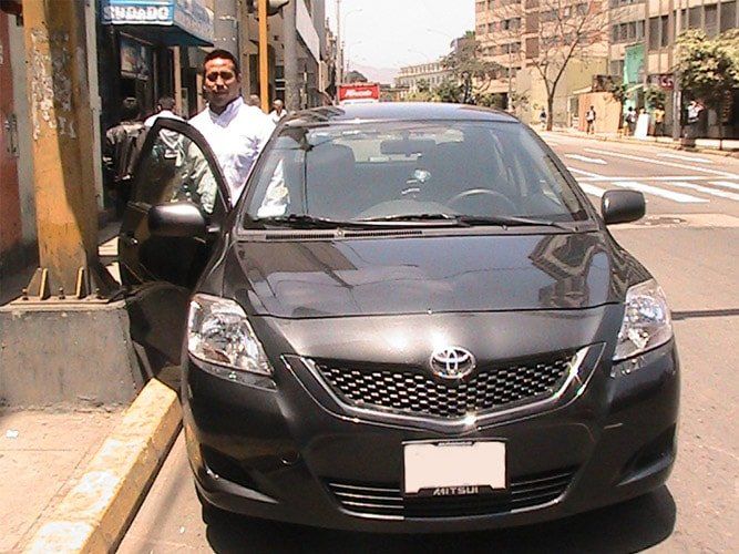 Hombre de pie junto a un sedán Toyota negro estacionado en una calle de la ciudad.