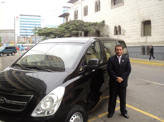 Hombre con traje oscuro de pie junto a una furgoneta negra en una calle de la ciudad cerca de un gran edificio.