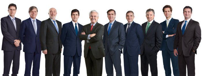 Nueve profesionales de negocios vestidos con trajes oscuros posan uno al lado del otro sobre un fondo blanco.