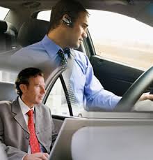 Dos hombres vestidos de traje dentro de un coche, uno al volante y el otro sentado en el asiento trasero.