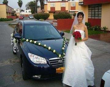 Novia con vestido blanco sosteniendo un ramo rojo, de pie junto a un coche negro decorado en una calle residencial.