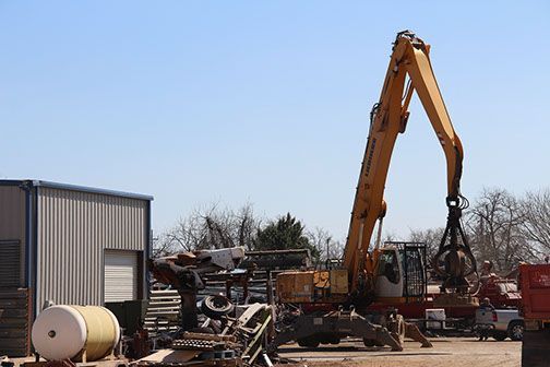 Large yellow excavator at an outdoor construction yard beside a metal building and equipment.