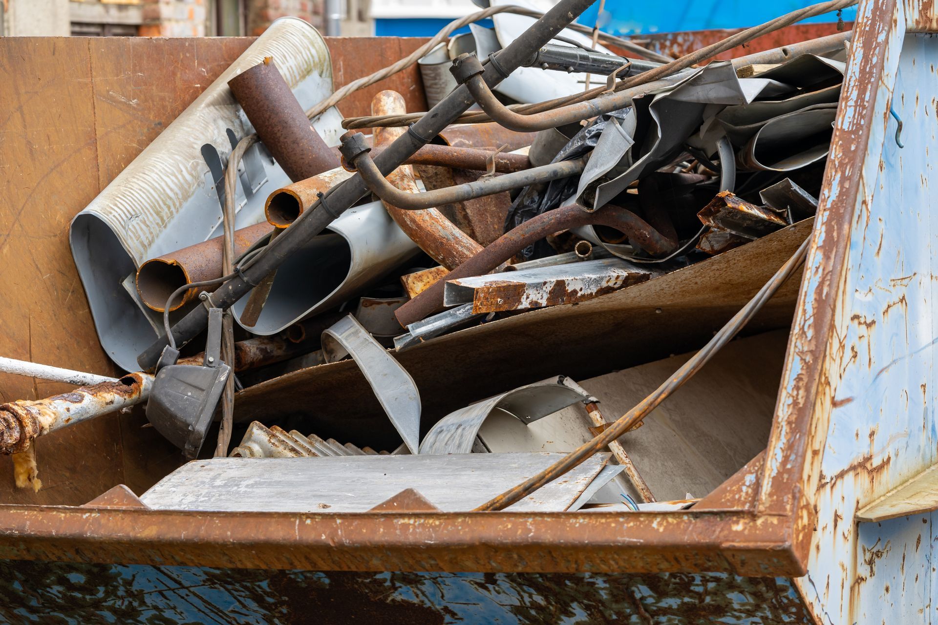 A rusty dumpster filled with bent metal pipes and scrap in an outdoor industrial setting.