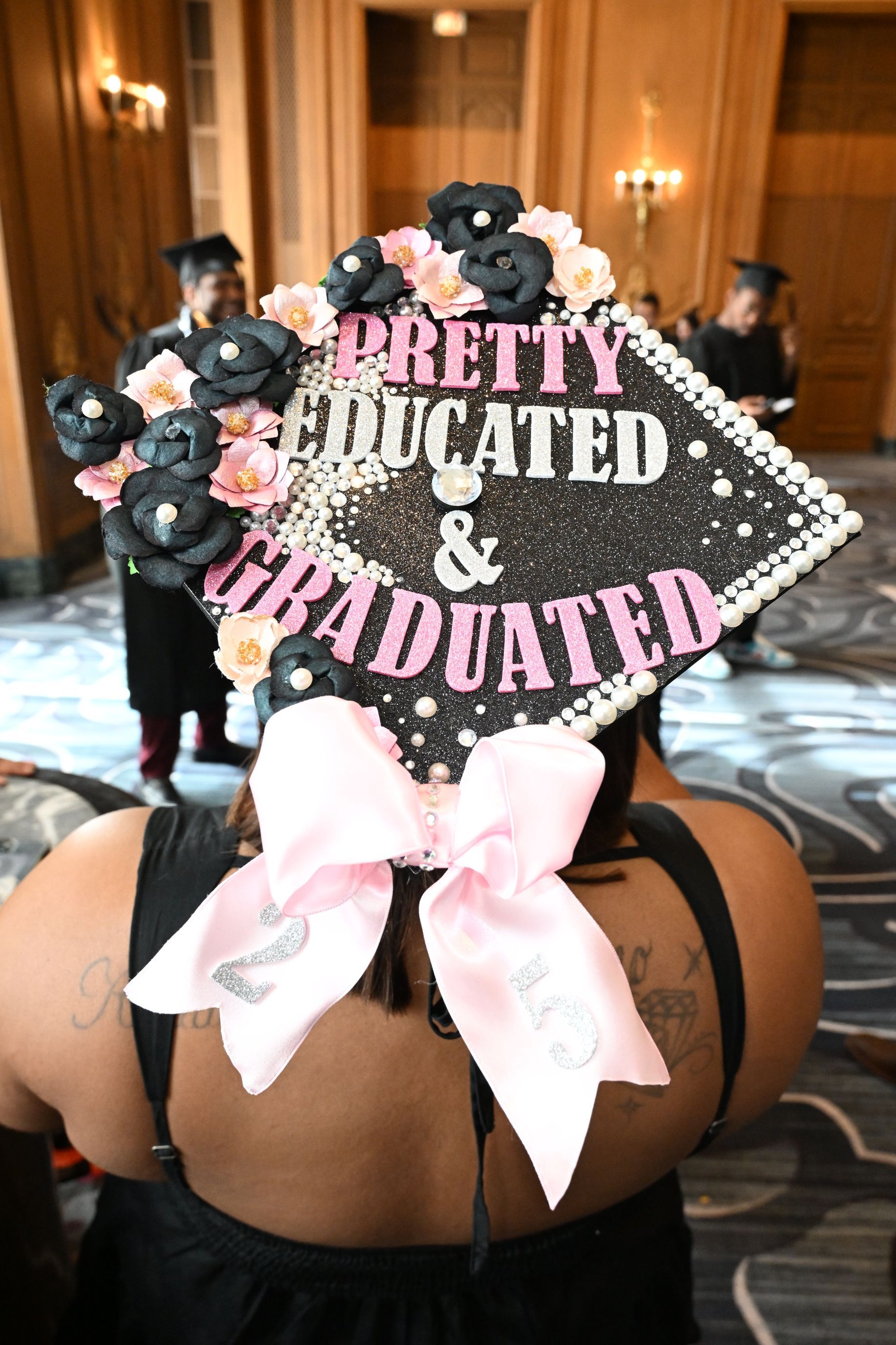 Graduate wearing a decorated cap with “Pretty Educated & Graduated.” Pink bow, black flowers, and pearls.