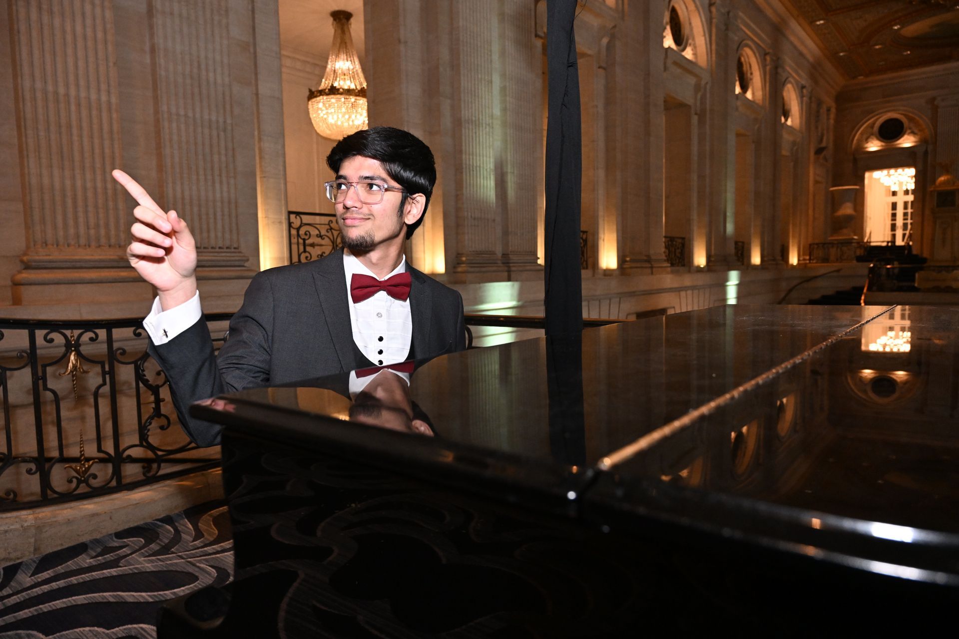 Man in suit with a bow tie pointing upward near a grand piano in a formal hall with columns and a chandelier.
