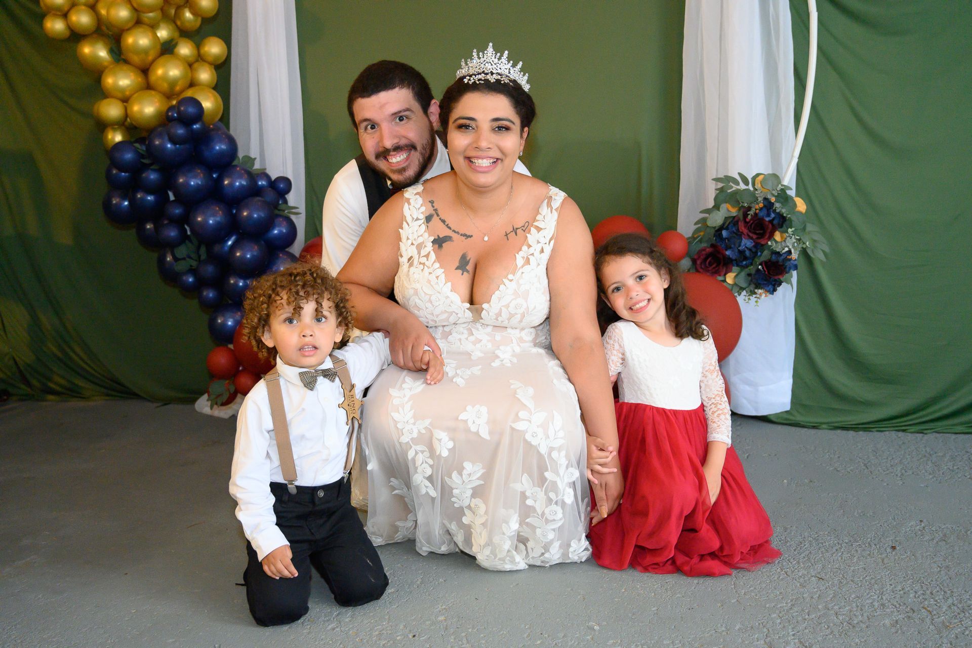 Family in wedding attire posing for photo. Bride in white dress, groom in vest, and two children smiling.