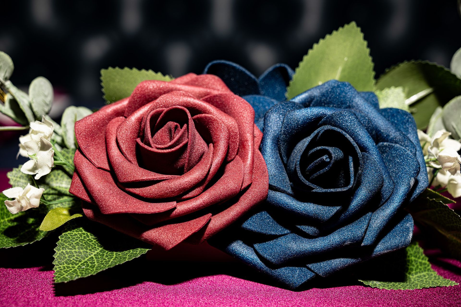 Two fabric roses, one red, one blue, with green leaves and white flowers, on a magenta surface.