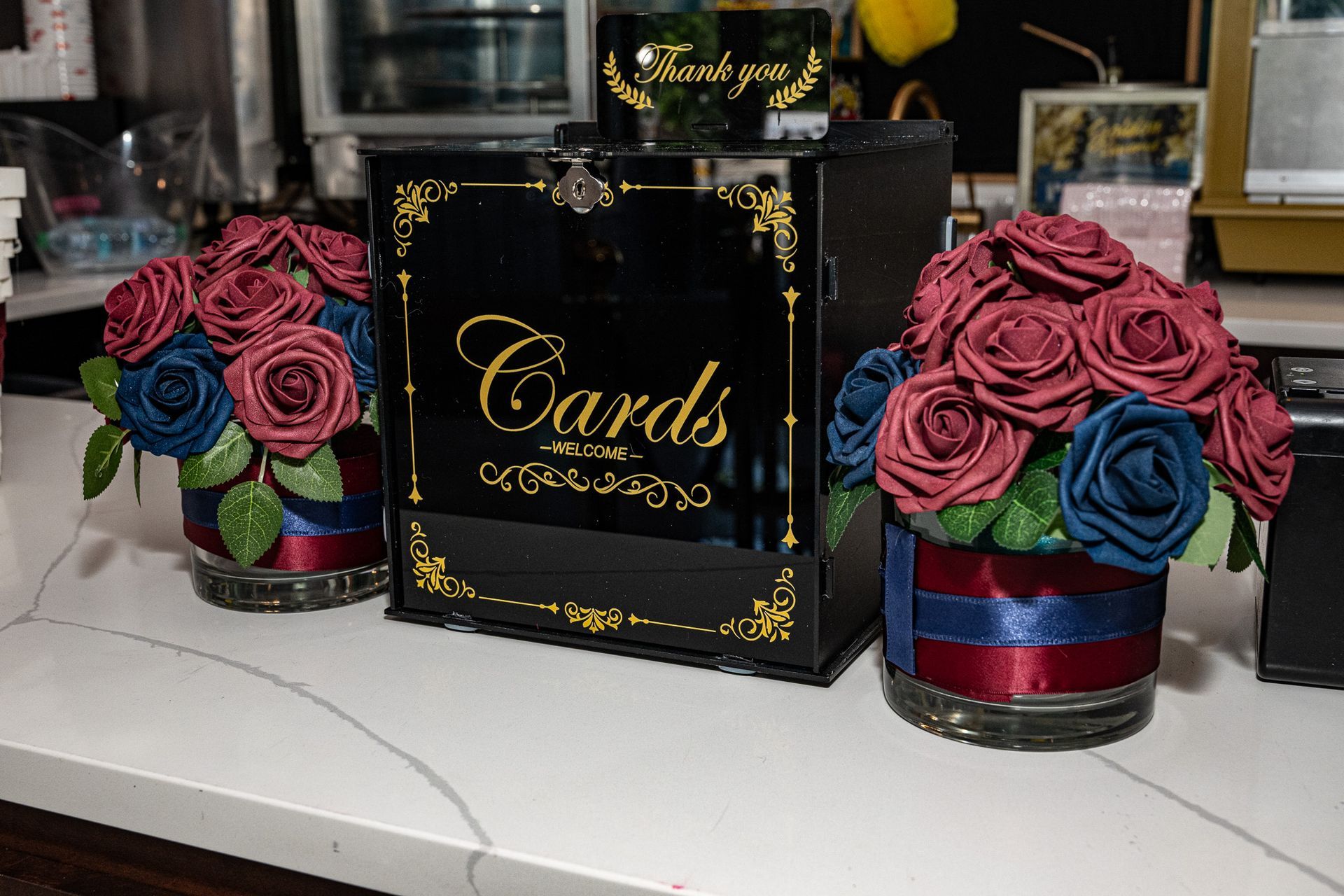 Black card box with gold lettering and decorative roses in vases, on a counter.