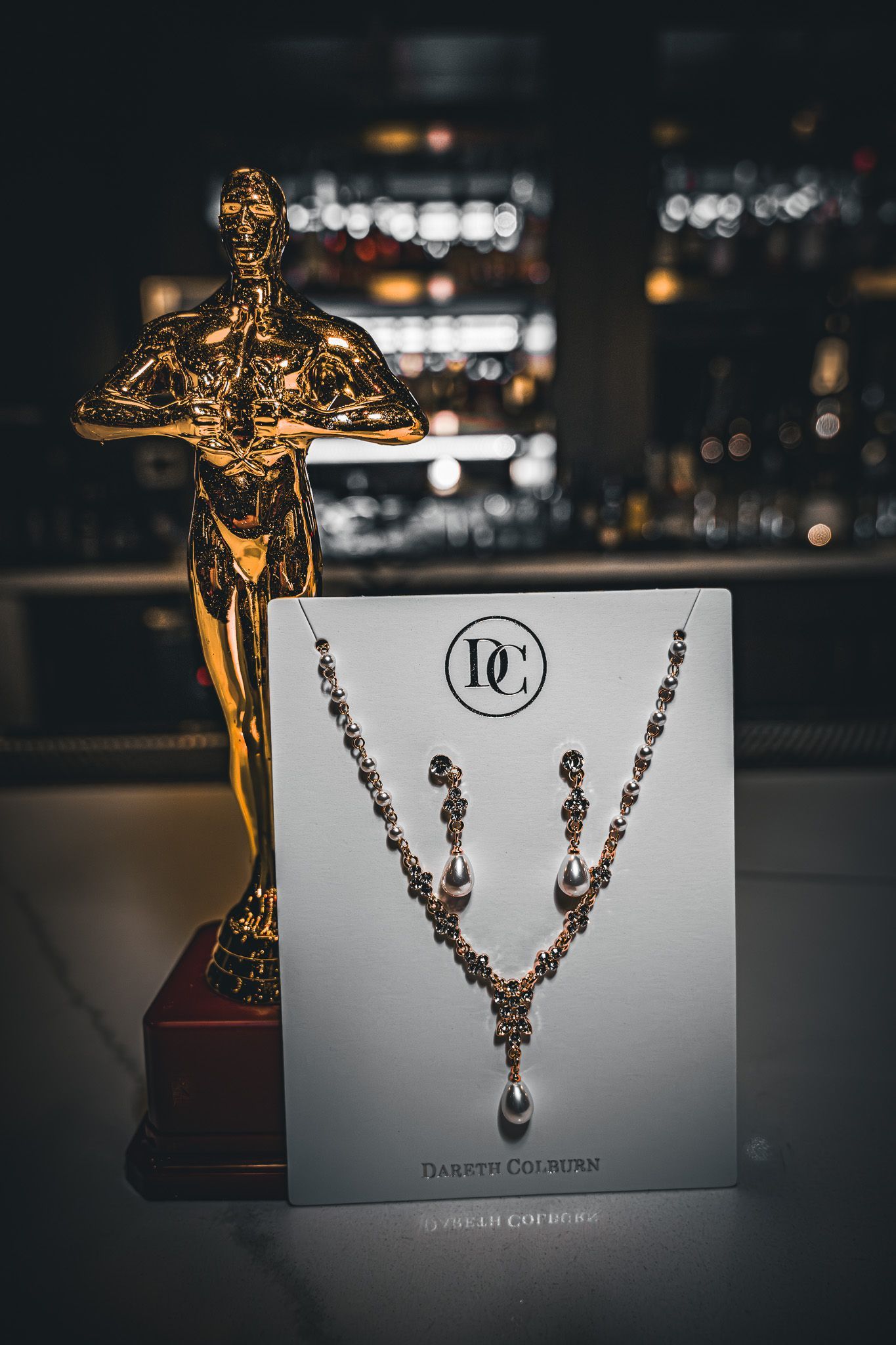 Golden necklace and earrings on display card with Oscar statue in front of a bar.