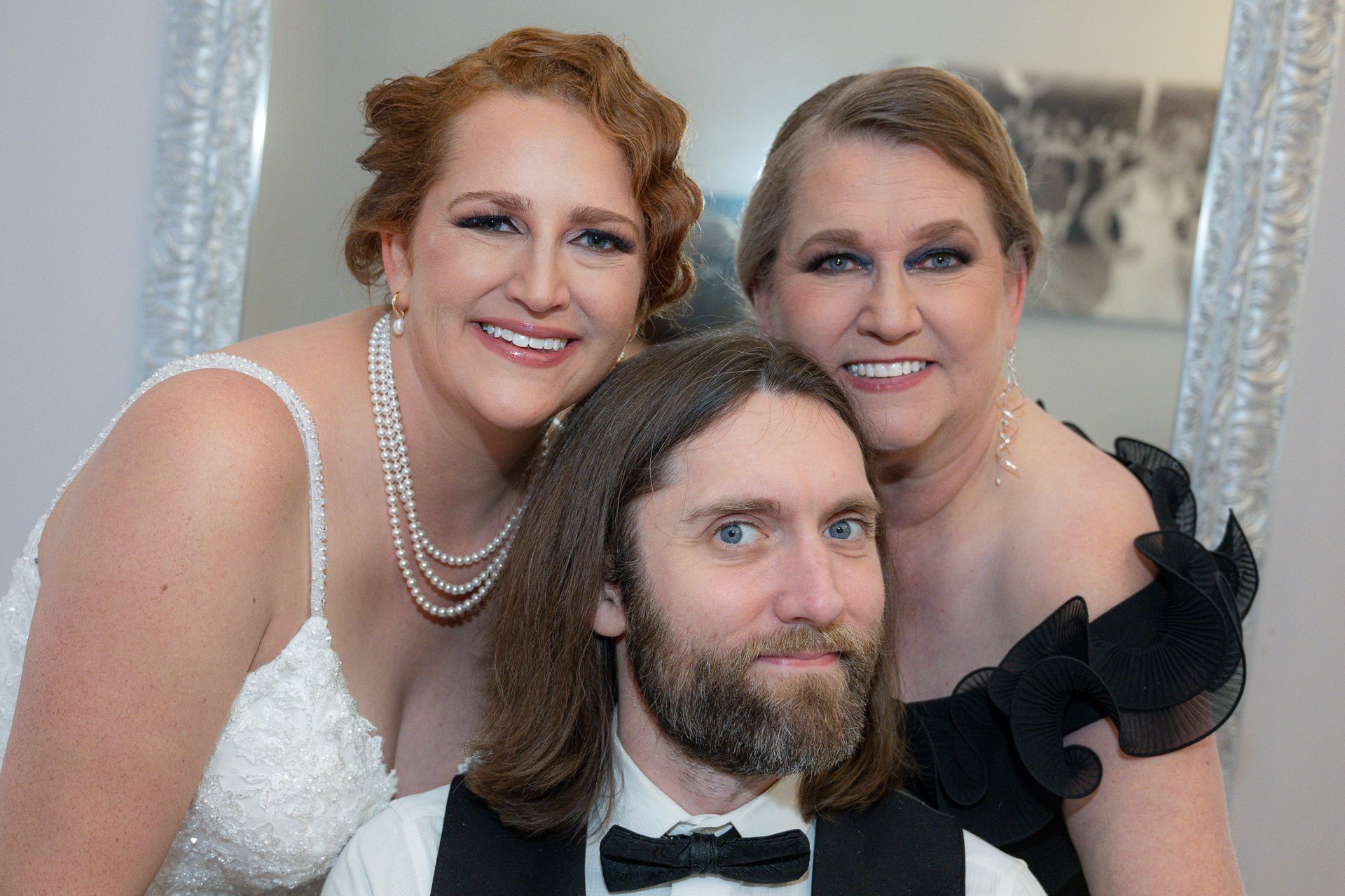 A man in a tuxedo smiles between two women in formal attire; one wearing a white dress, the other in black.