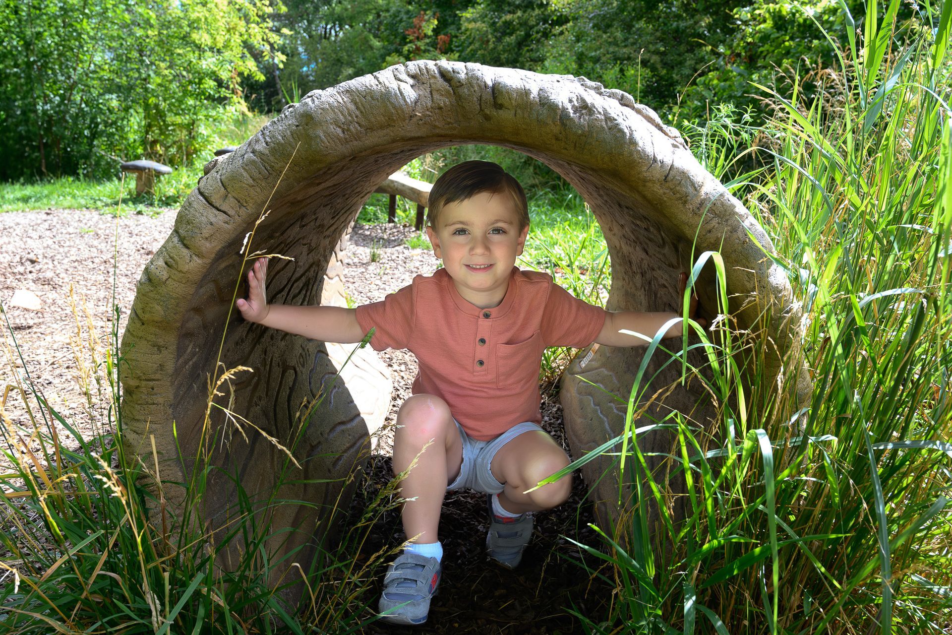 Young child crouches in a wooden tunnel, smiling. Green grass and foliage surround.