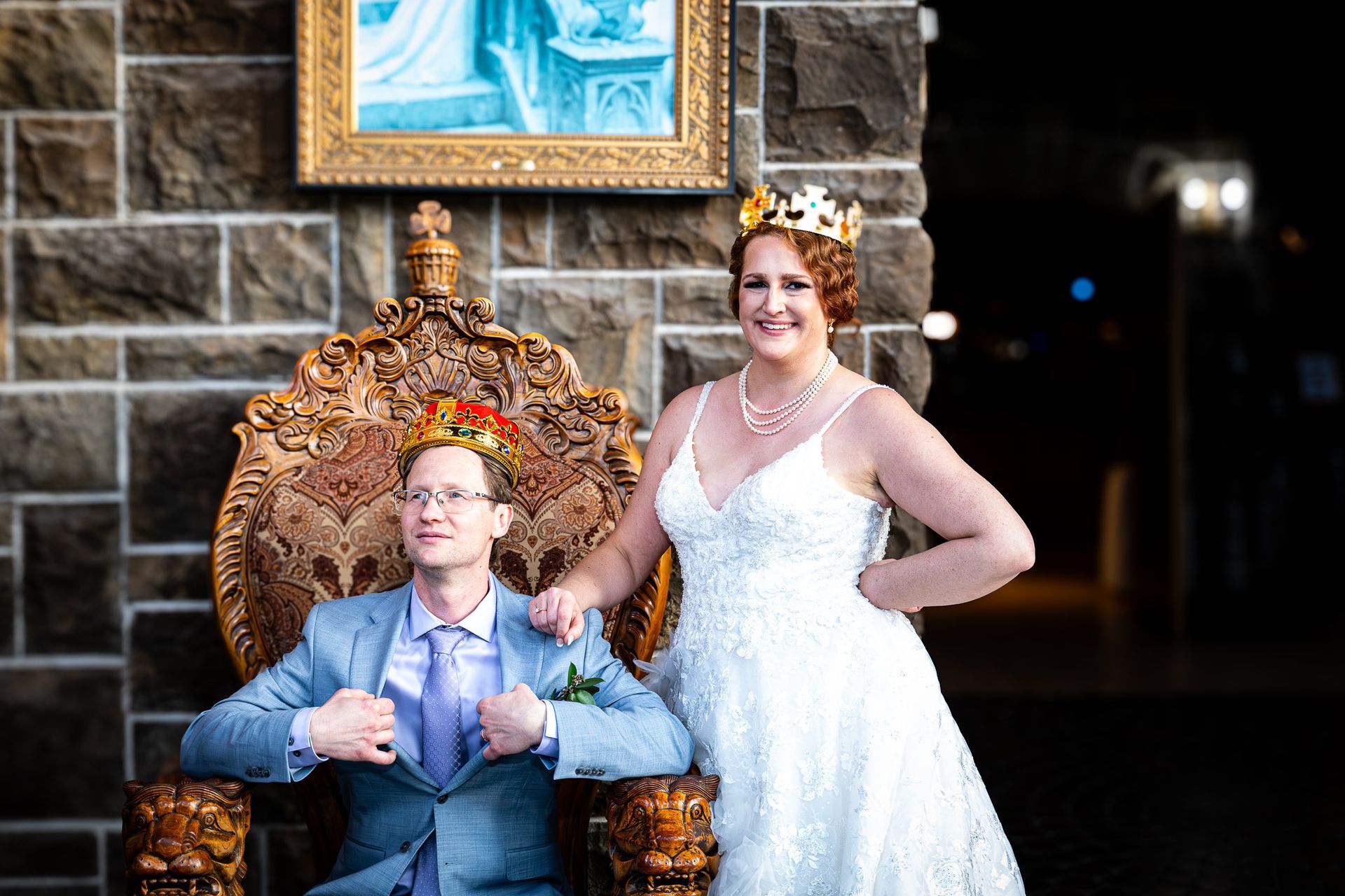 Bride and groom wearing crowns pose on a golden throne in front of a stone wall.