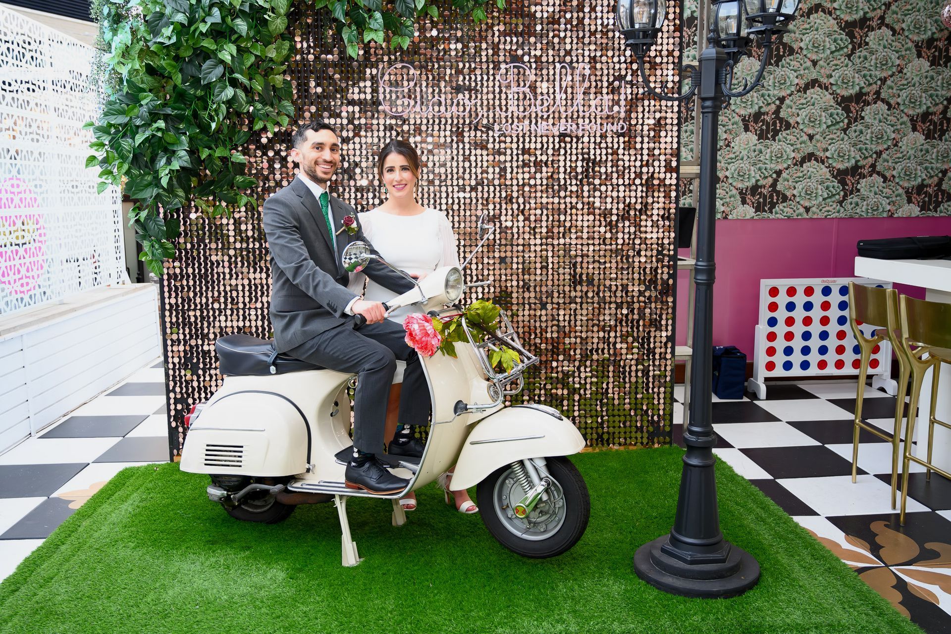 Couple posing on a cream scooter in front of a sequined backdrop; surrounded by greenery and a lamppost.