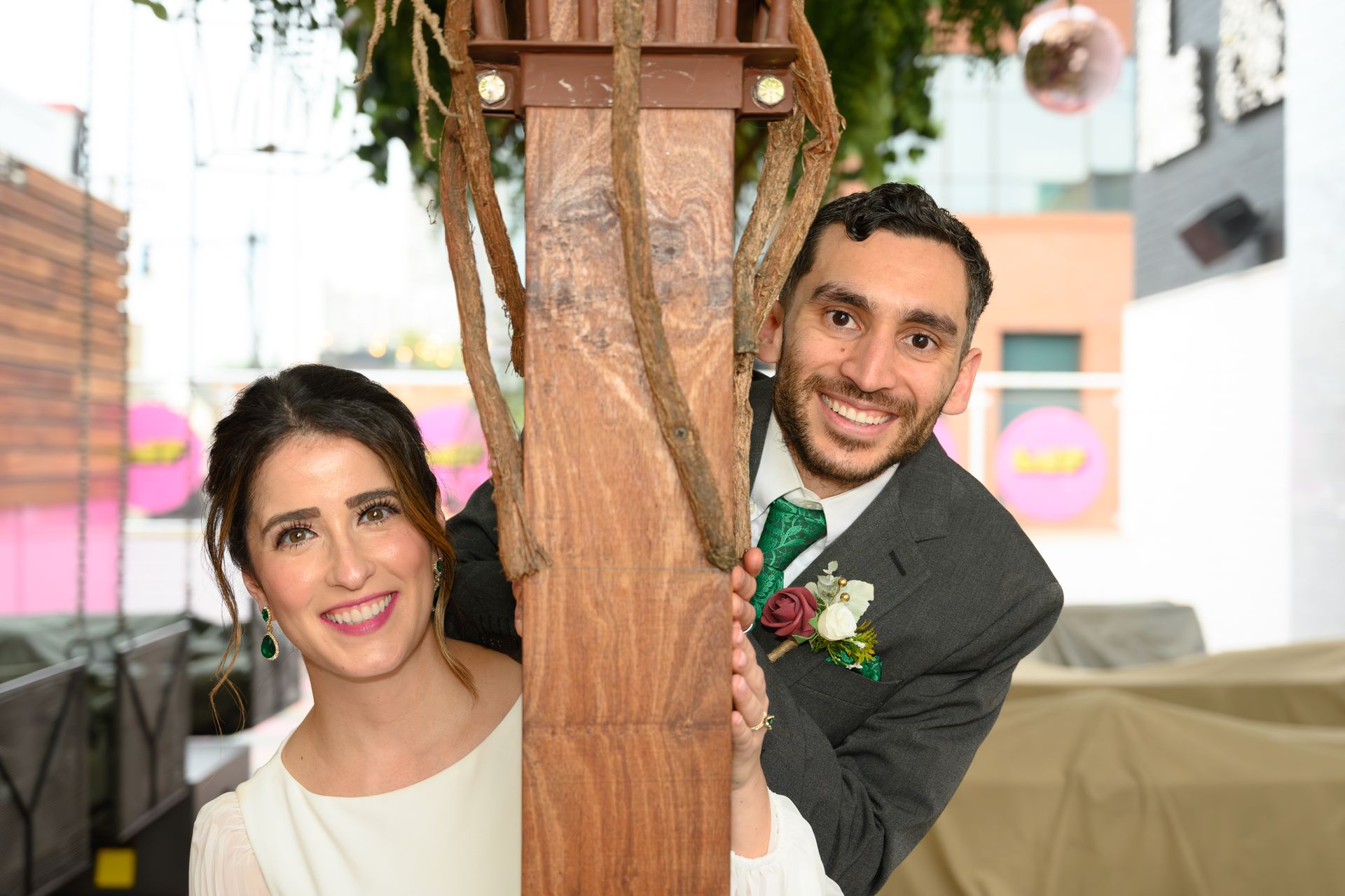 Couple smiling, peering around a wooden post decorated with vines, likely at a wedding.