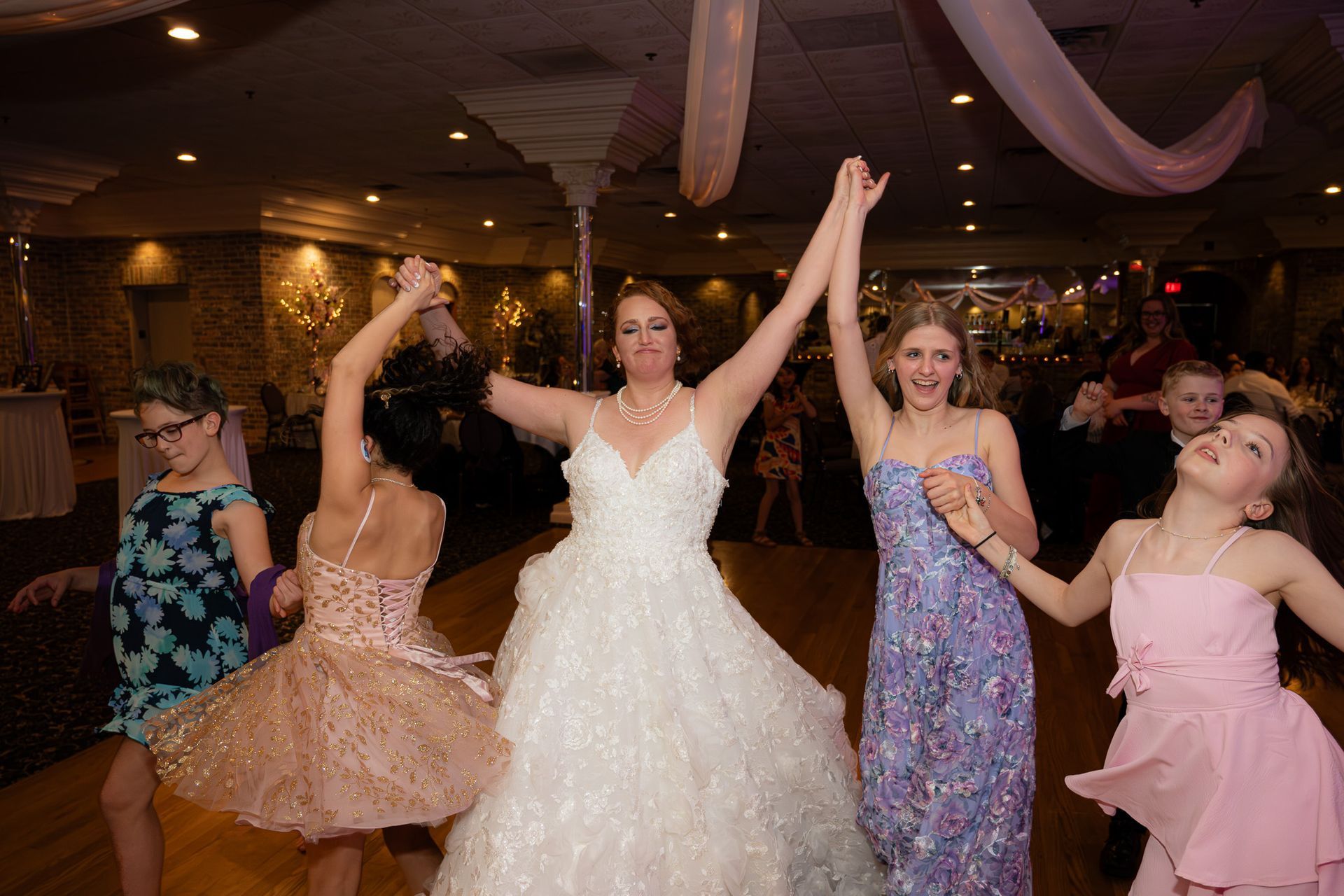 Bride dancing with young girls, holding hands, in a decorated ballroom.