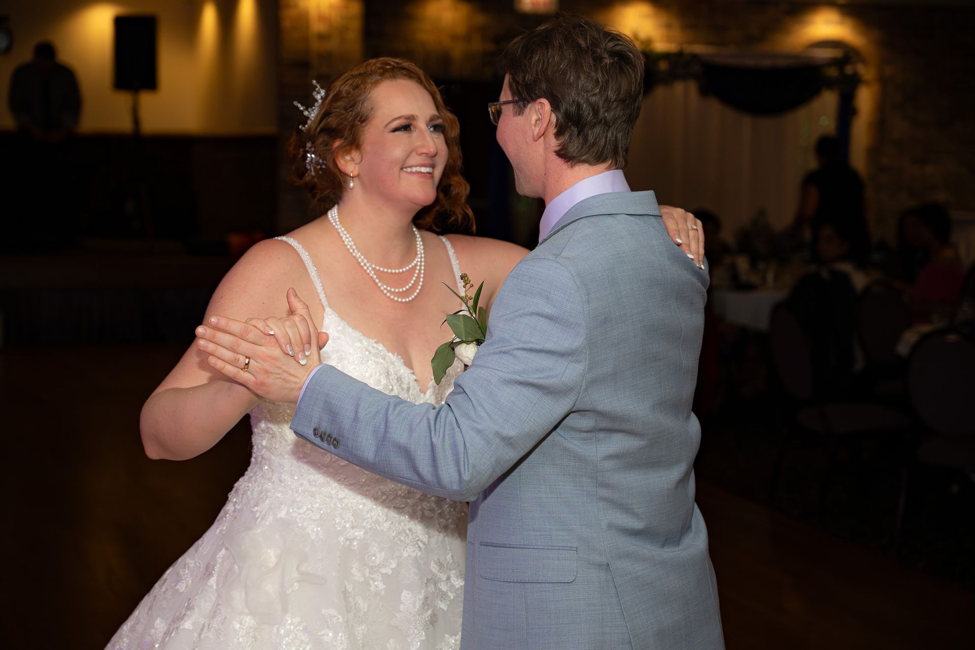 Bride and groom dance at reception; bride in white gown, groom in light blue suit; smiling, indoors.