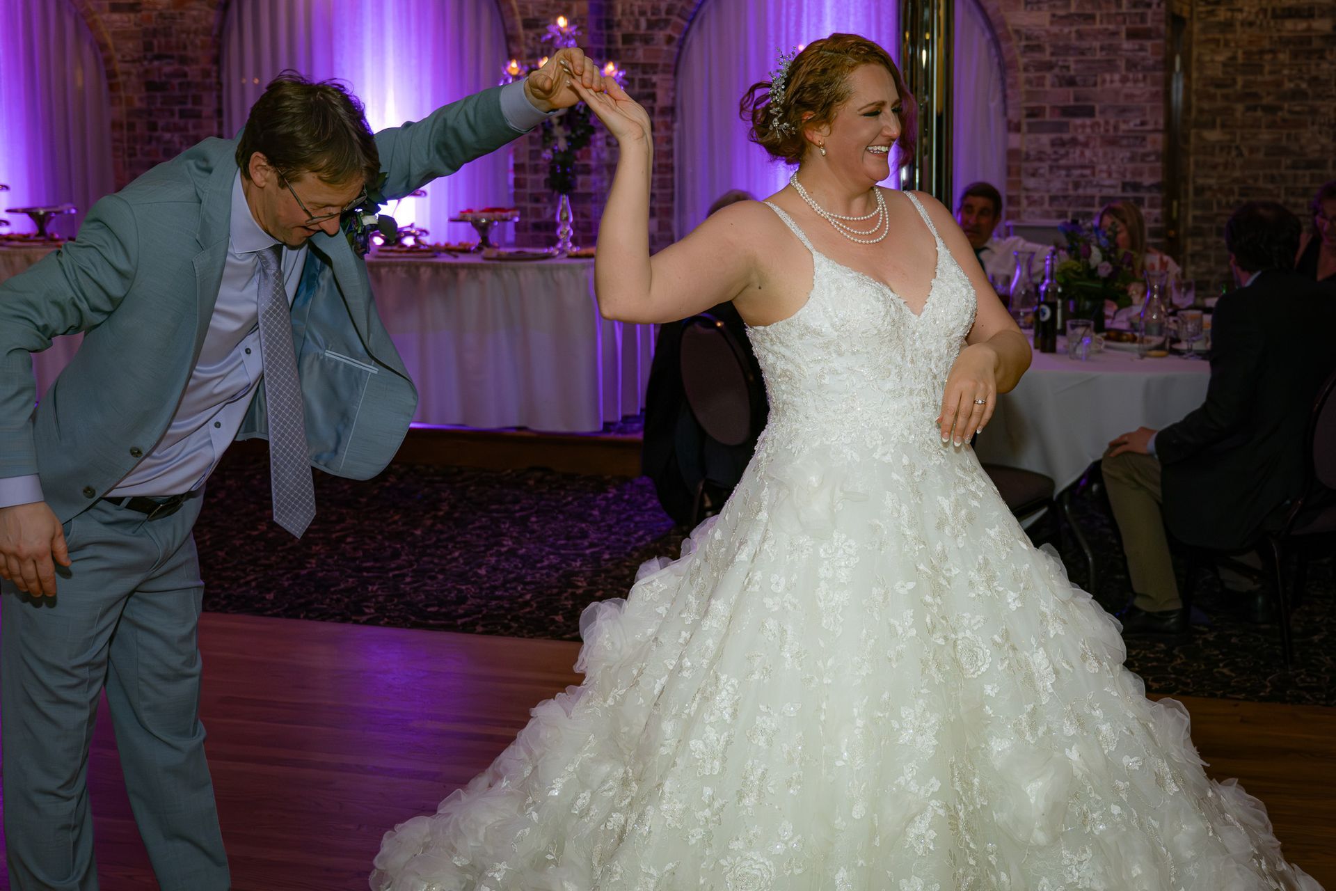 Bride in white gown dancing with a man in a blue suit at a wedding reception.