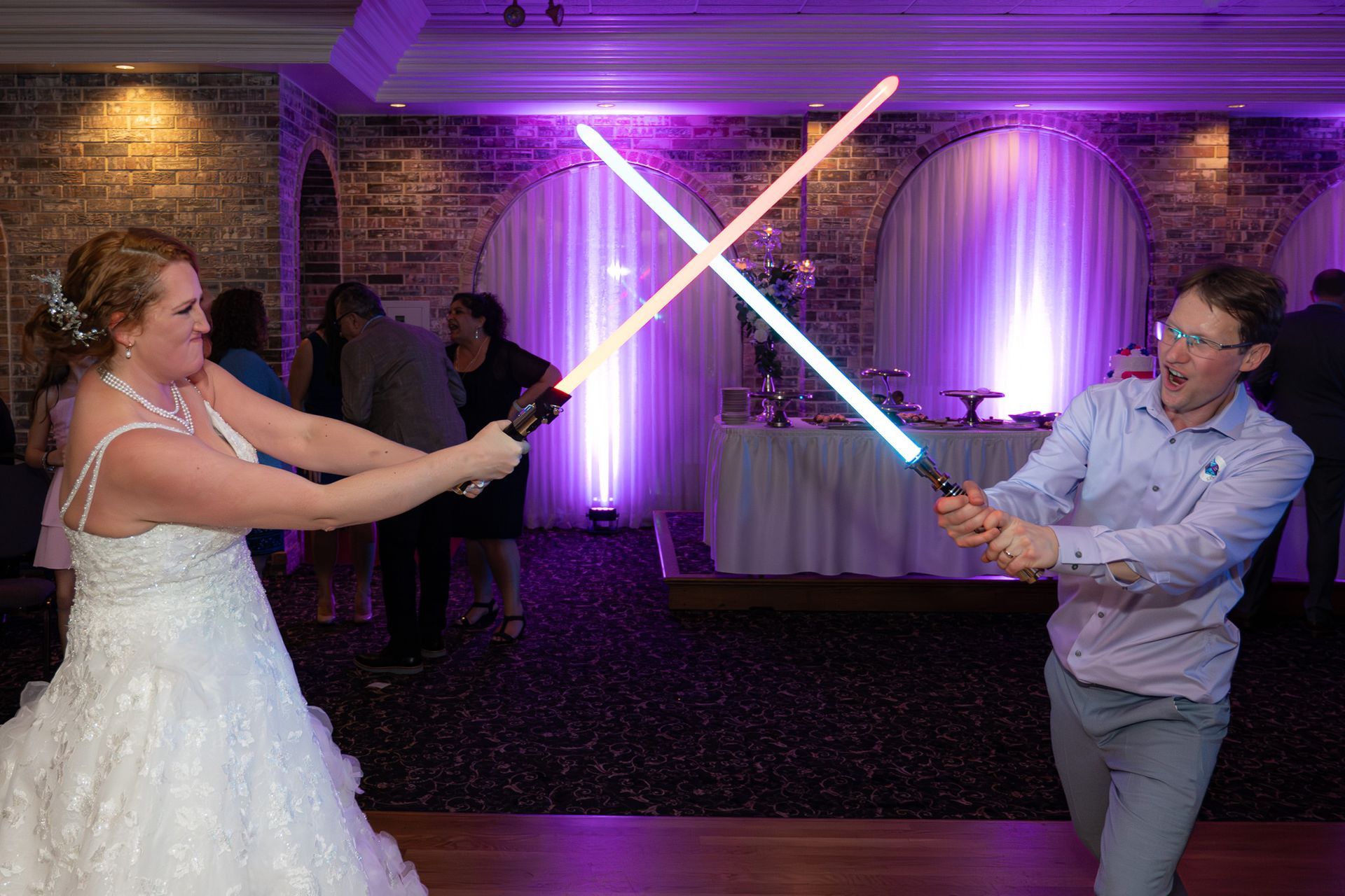 Bride and groom holding lightsabers in a wedding reception; fighting playfully with glowing sabers.