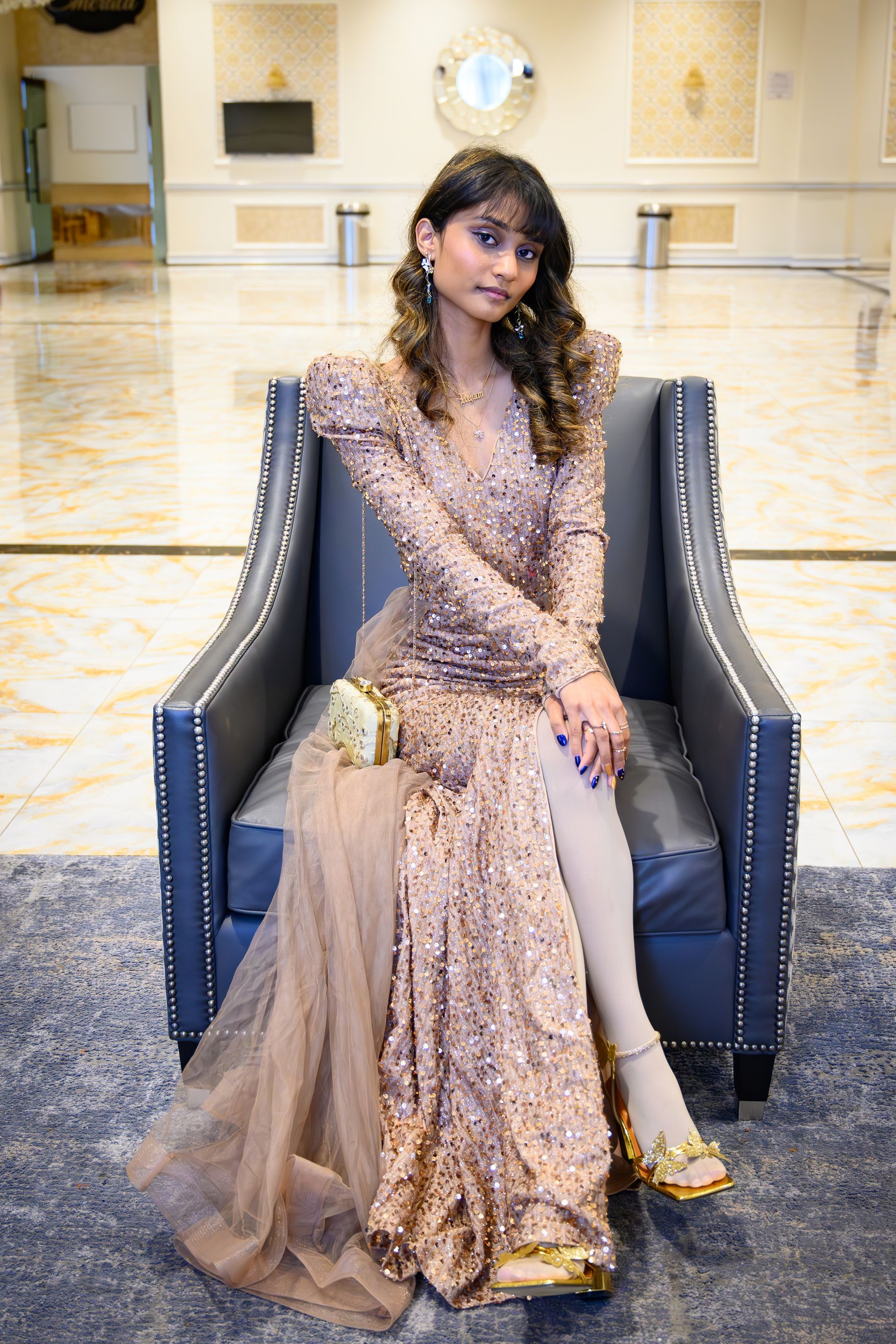Woman in a sparkly gown sits in a dark blue chair with gold sandals and a clutch in a hotel lobby.