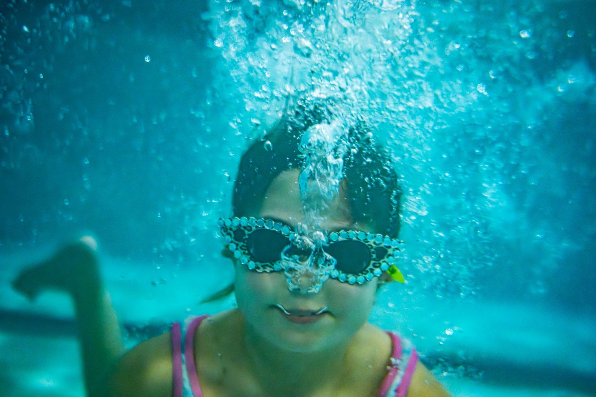 Girl underwater, wearing goggles, blowing bubbles in a pool.