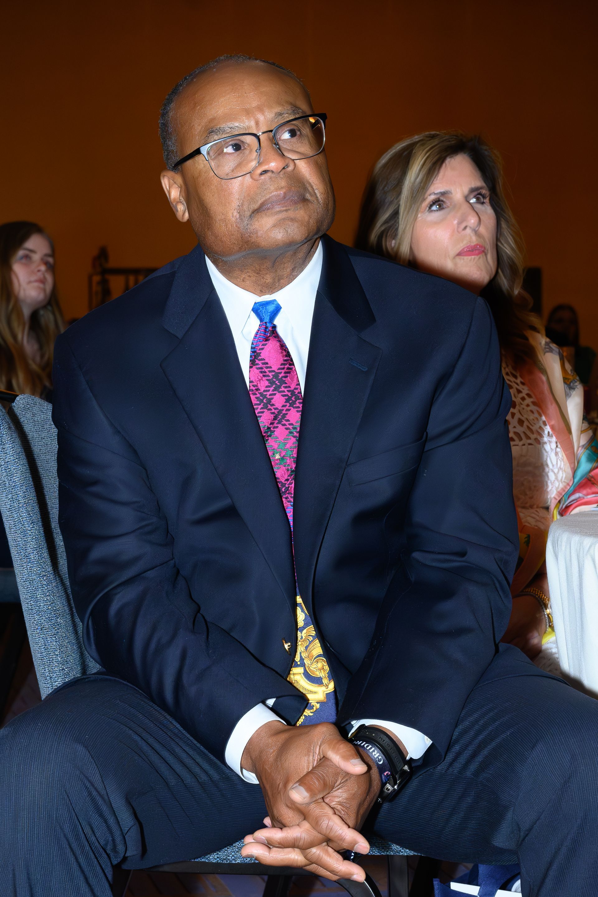 Man in suit and tie seated, looking up with a contemplative expression. Woman to the right.