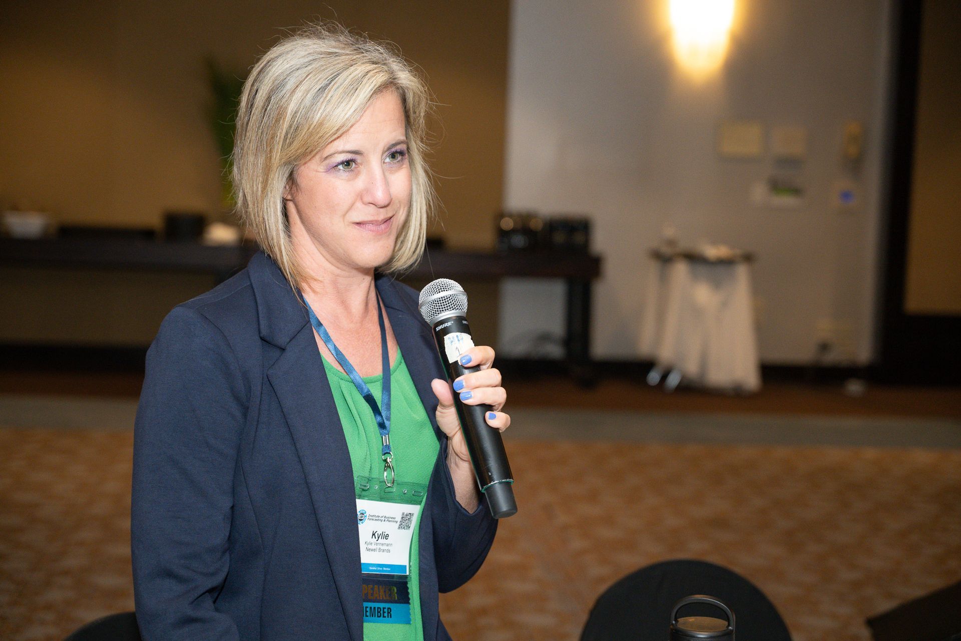 Woman in a blazer and green shirt speaking into a microphone at an event, smiling.