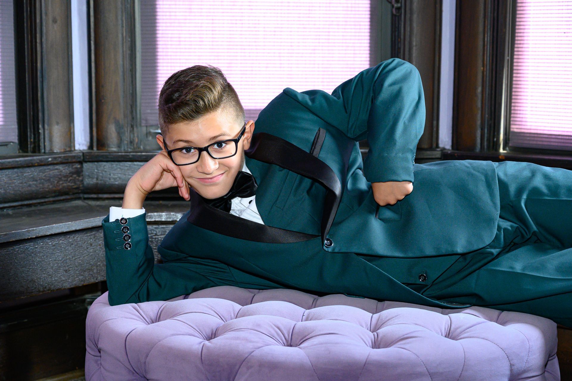 Boy wearing glasses and a teal suit, reclining on a purple ottoman, smiling.