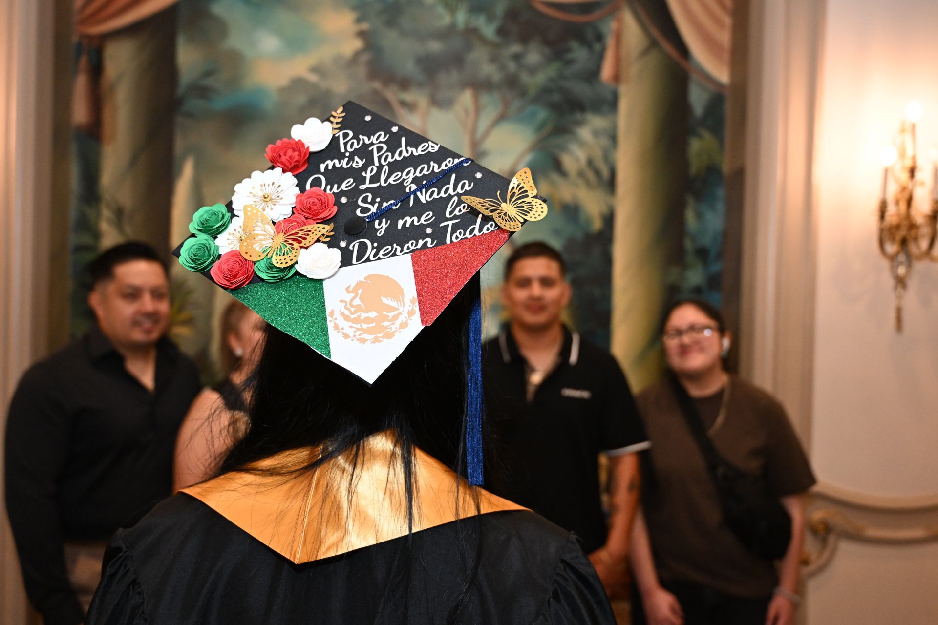 Person in graduation gown with decorated cap; family in background, interior setting.