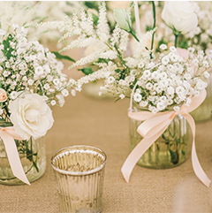White flowers in glass jars tied with pink ribbons on a burlap tablecloth with a votive candle.