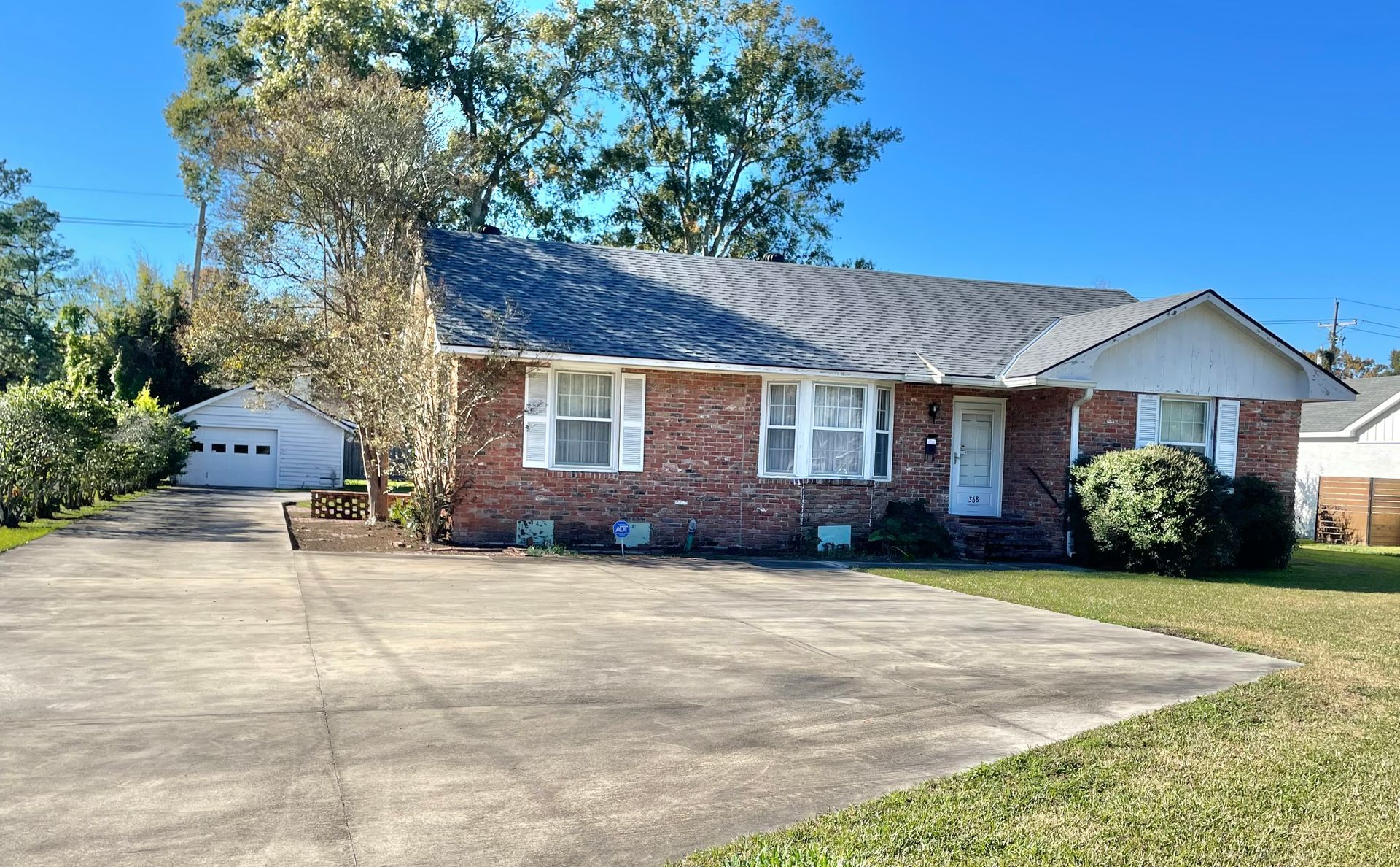 Brick ranch house with attached carport, wide concrete driveway, and trees under a clear blue sky
