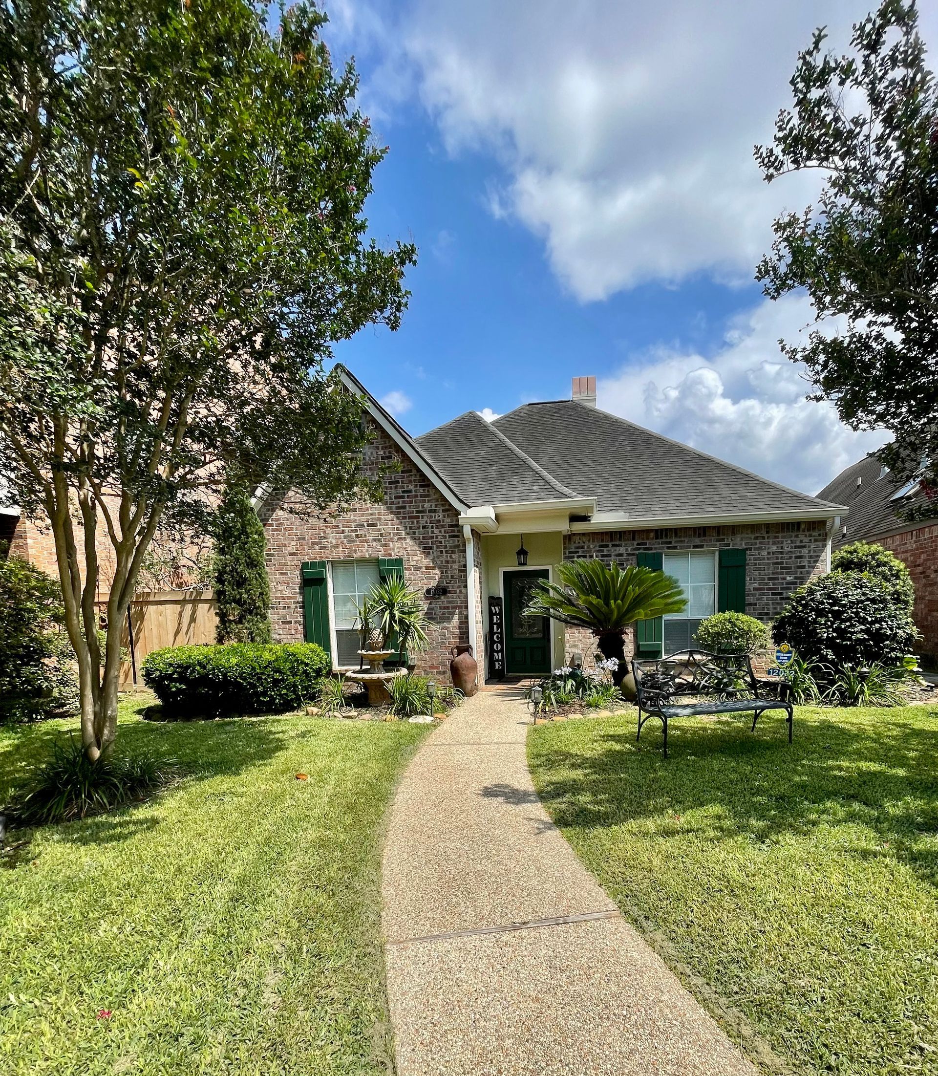 Single-story brick house with a gravel path, green lawn, and trees under a partly cloudy sky