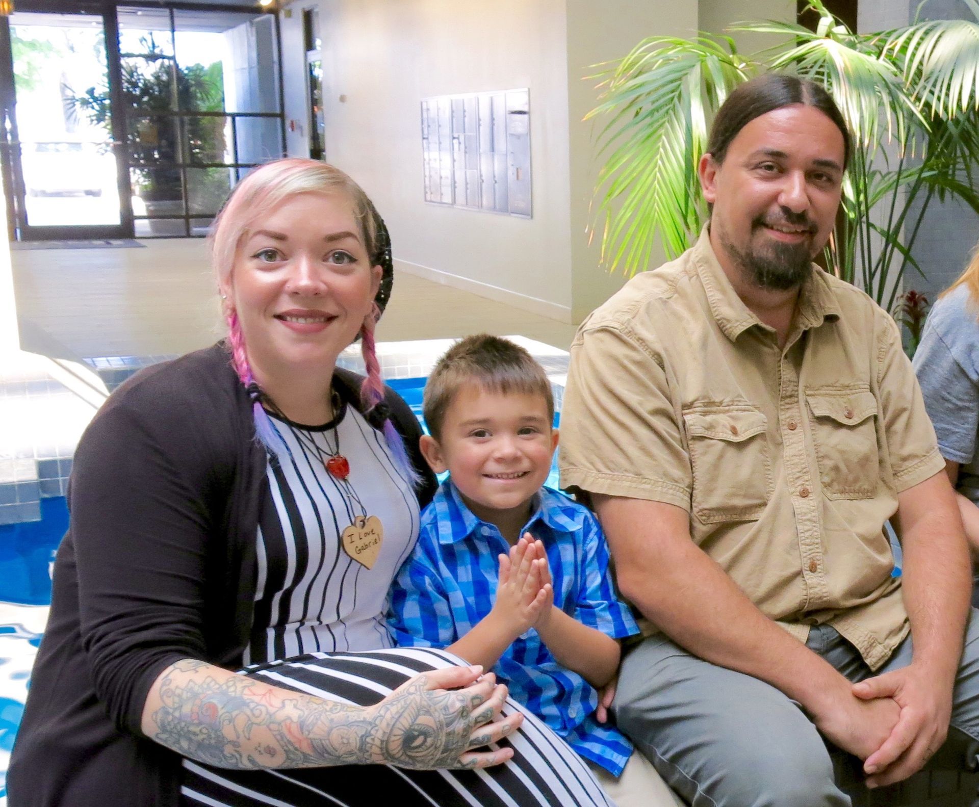 Three people smiling on a couch in a bright lobby, with a plant and window behind them.