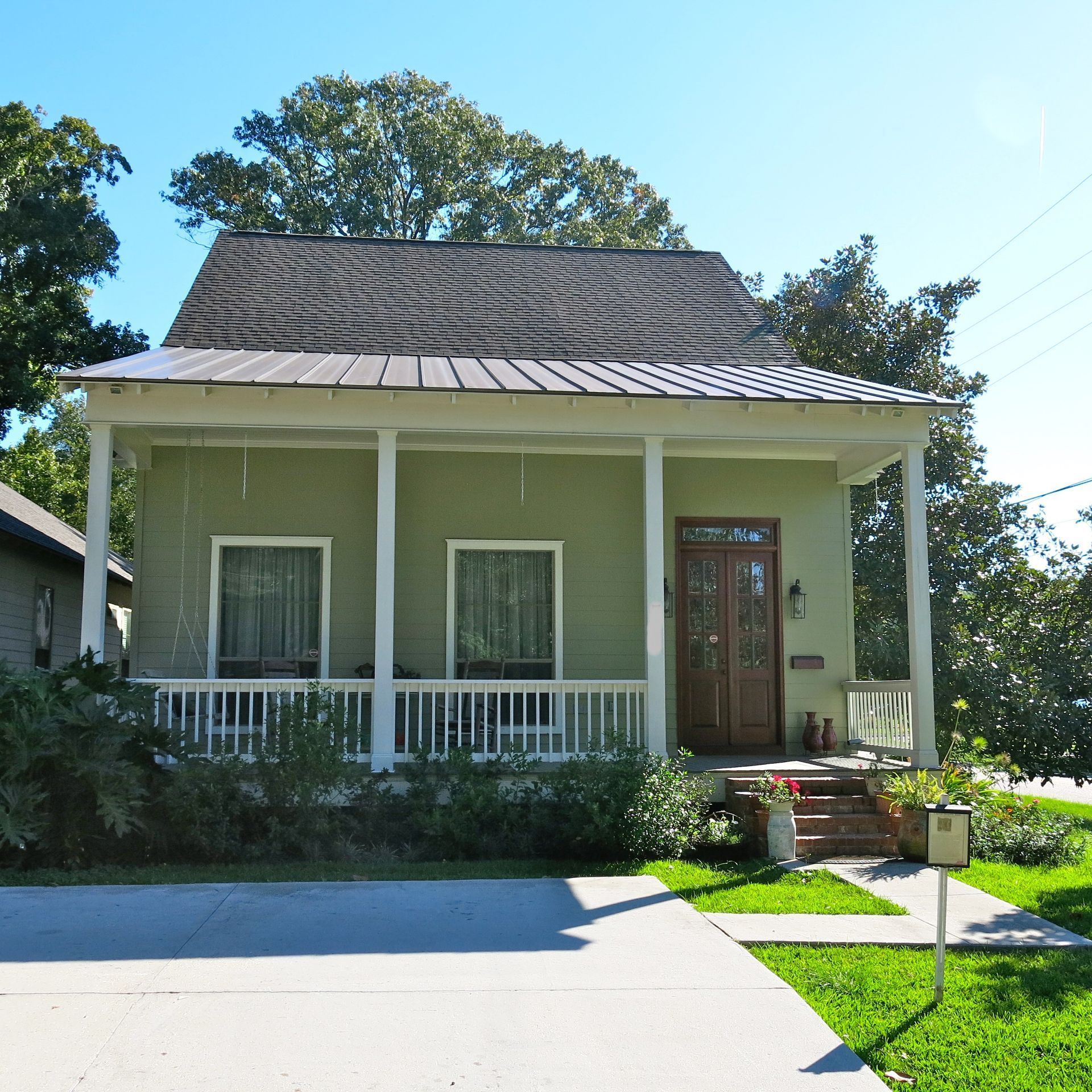 Small green bungalow with a front porch, white railing, and a driveway in bright sunlight.