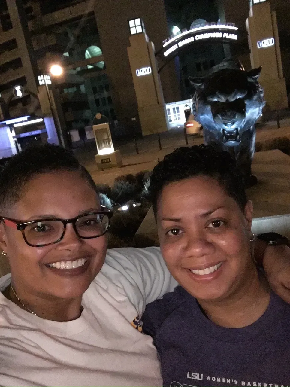Two smiling people posing for a selfie at night, with a lit fountain and buildings behind them