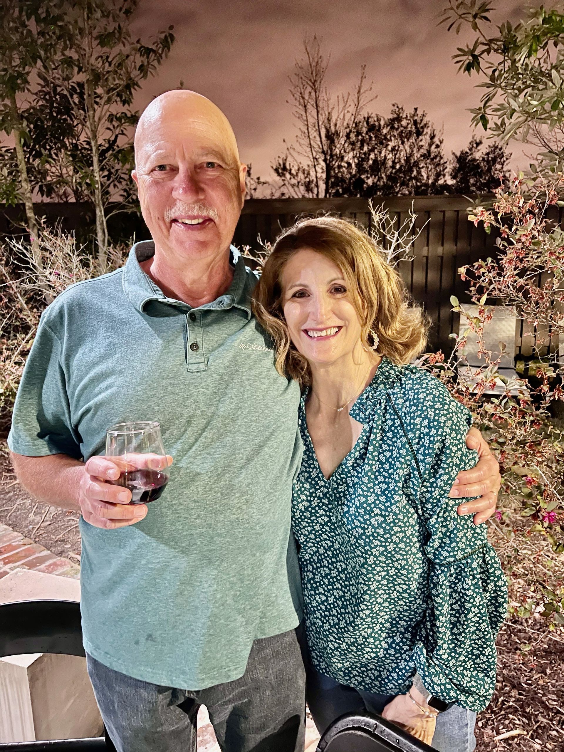 Smiling couple posing outdoors at night, man holding a drink, woman in teal blouse, festive lights behind them