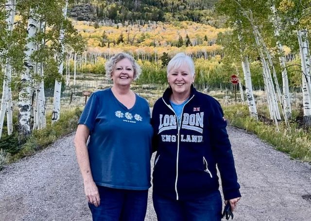 Two smiling people stand on a gravel path with white trees and a mountain in the background.