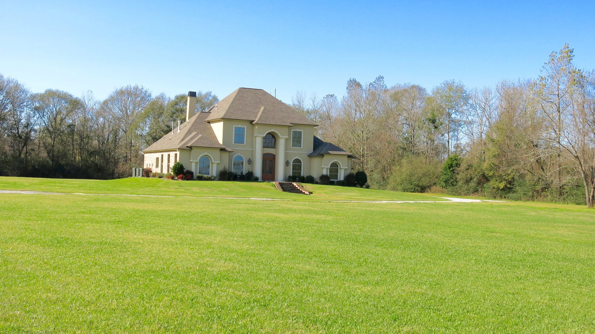 Large beige house with a tan roof on a wide green lawn, bordered by trees under a clear blue sky