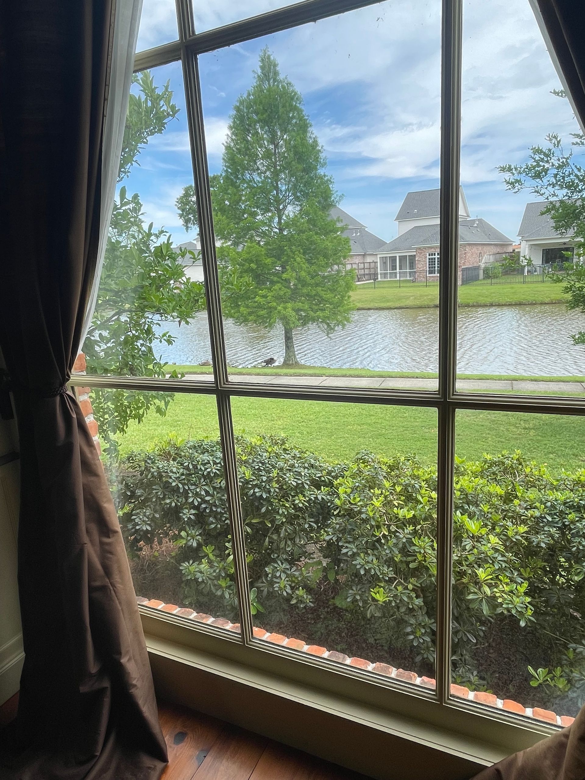 View through a window of a calm pond, hedge, and houses under a cloudy sky