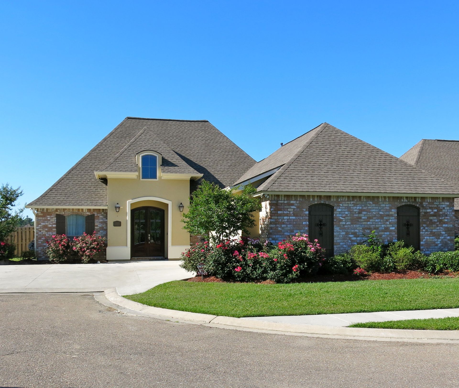 Large suburban house with stone and stucco exterior, arched entry, and landscaped flower beds under a clear blue sky