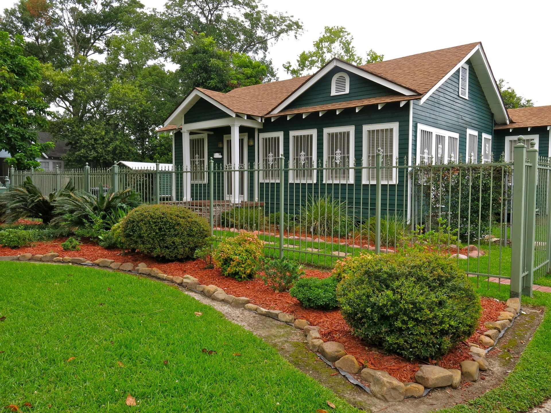Small green house with white trim and fenced yard, bordered by flowers and shrubs