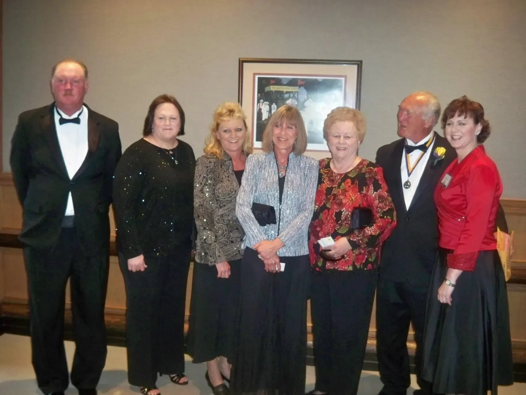Seven formally dressed people posing indoors at a banquet, with framed artwork on the wall.