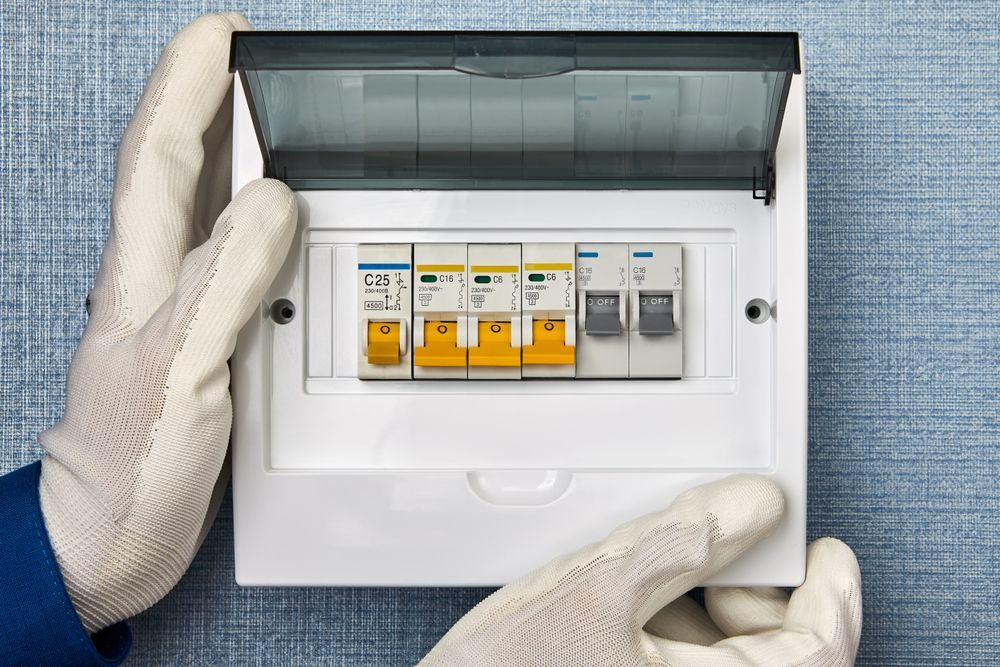 Hands in gloves holding an open circuit breaker box on a wall.