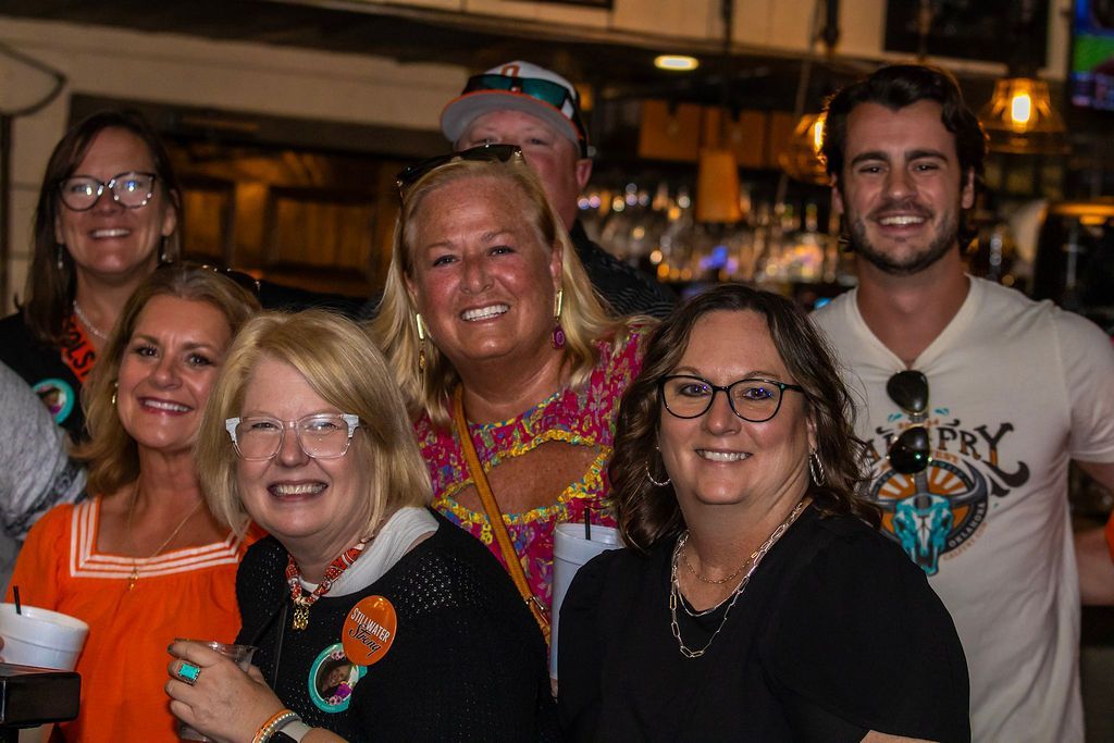 Group of smiling people pose indoors, possibly a bar; some wear glasses and colorful shirts.
