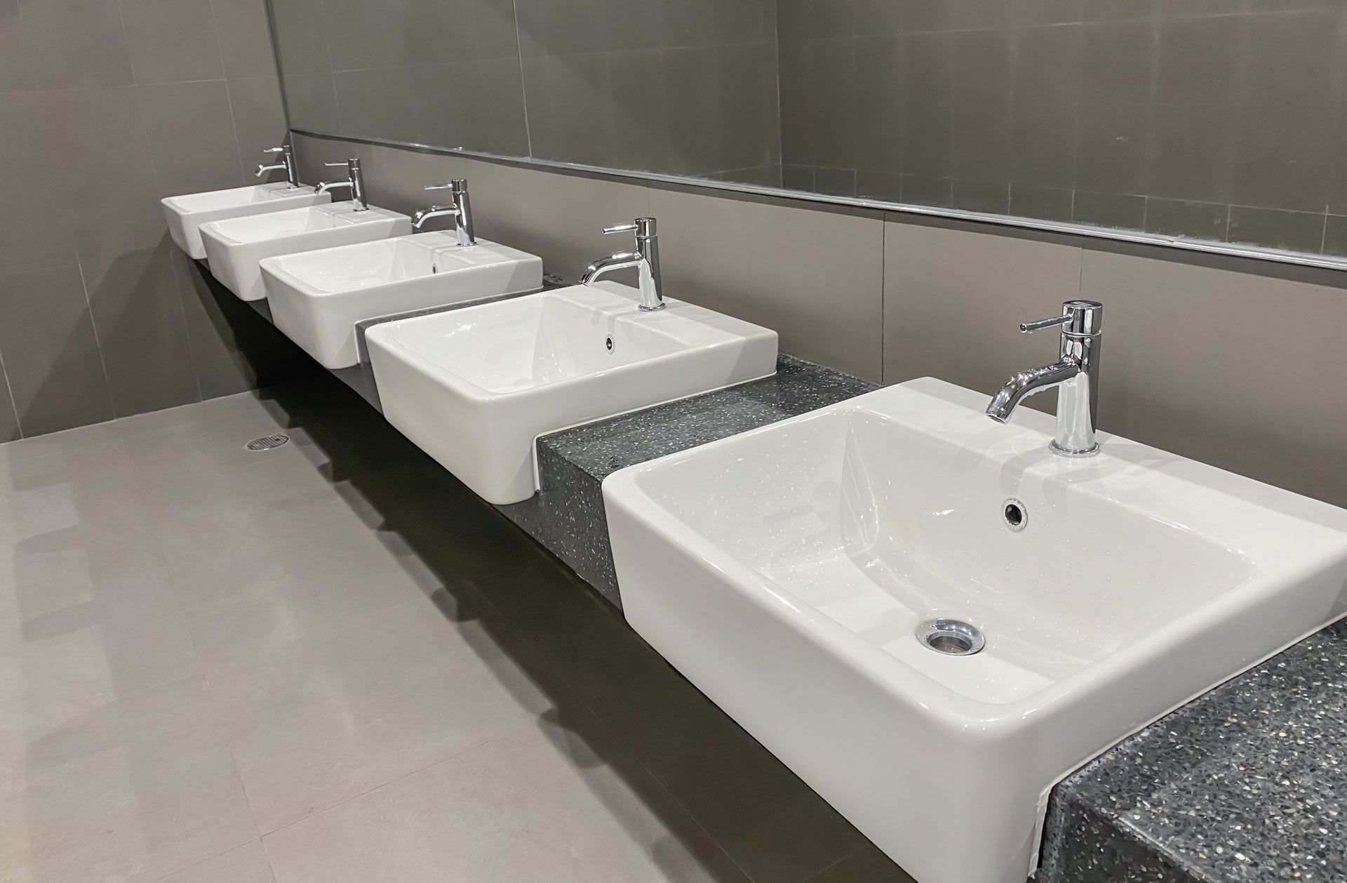 Row of white sinks with silver faucets along a gray tiled restroom wall