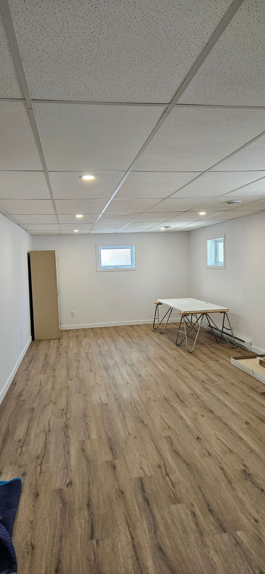 Une pièce avec un sol imitation bois marron clair, des murs blancs et un plafond blanc avec des spots encastrés. Une table est installée contre le mur du fond.