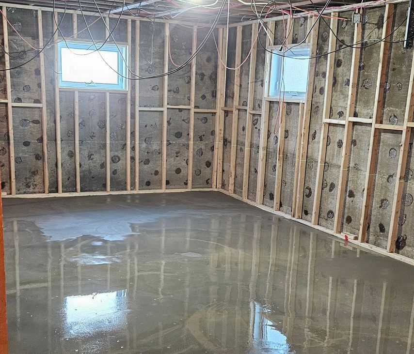 Intérieur d'un sous-sol en construction avec un plancher en béton fraîchement coulé et des murs isolés avec charpente apparente.