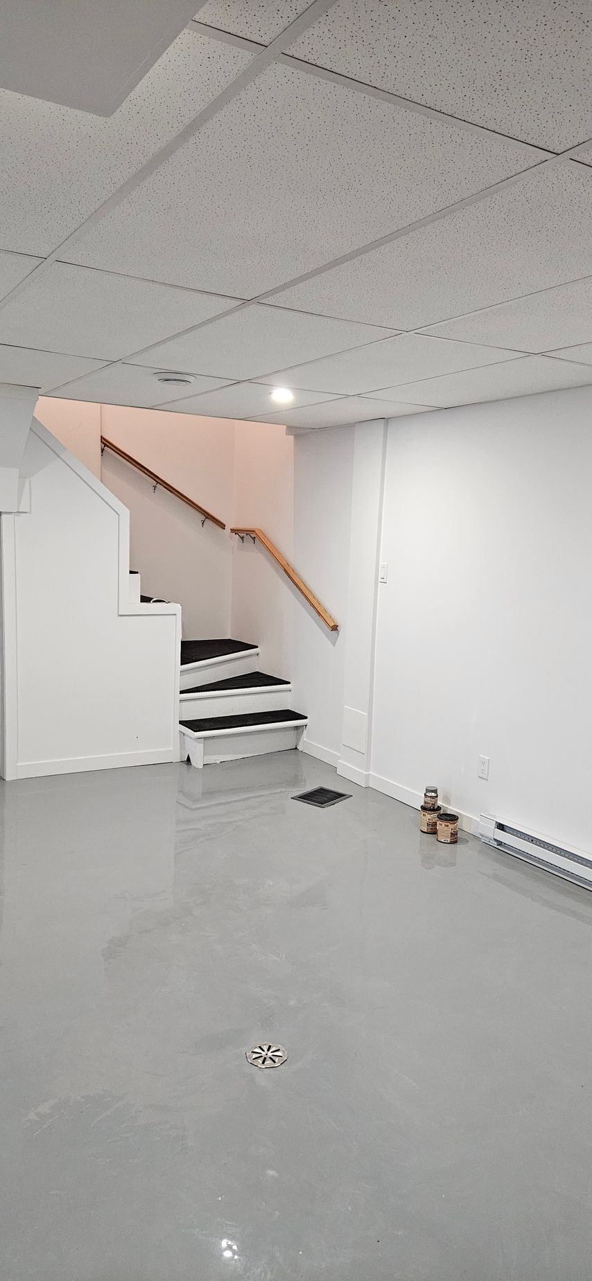 Un sous-sol vide aux murs blancs, au sol gris clair et à l'escalier avec rampe en bois. Le plafond est carrelé et un luminaire encastré.