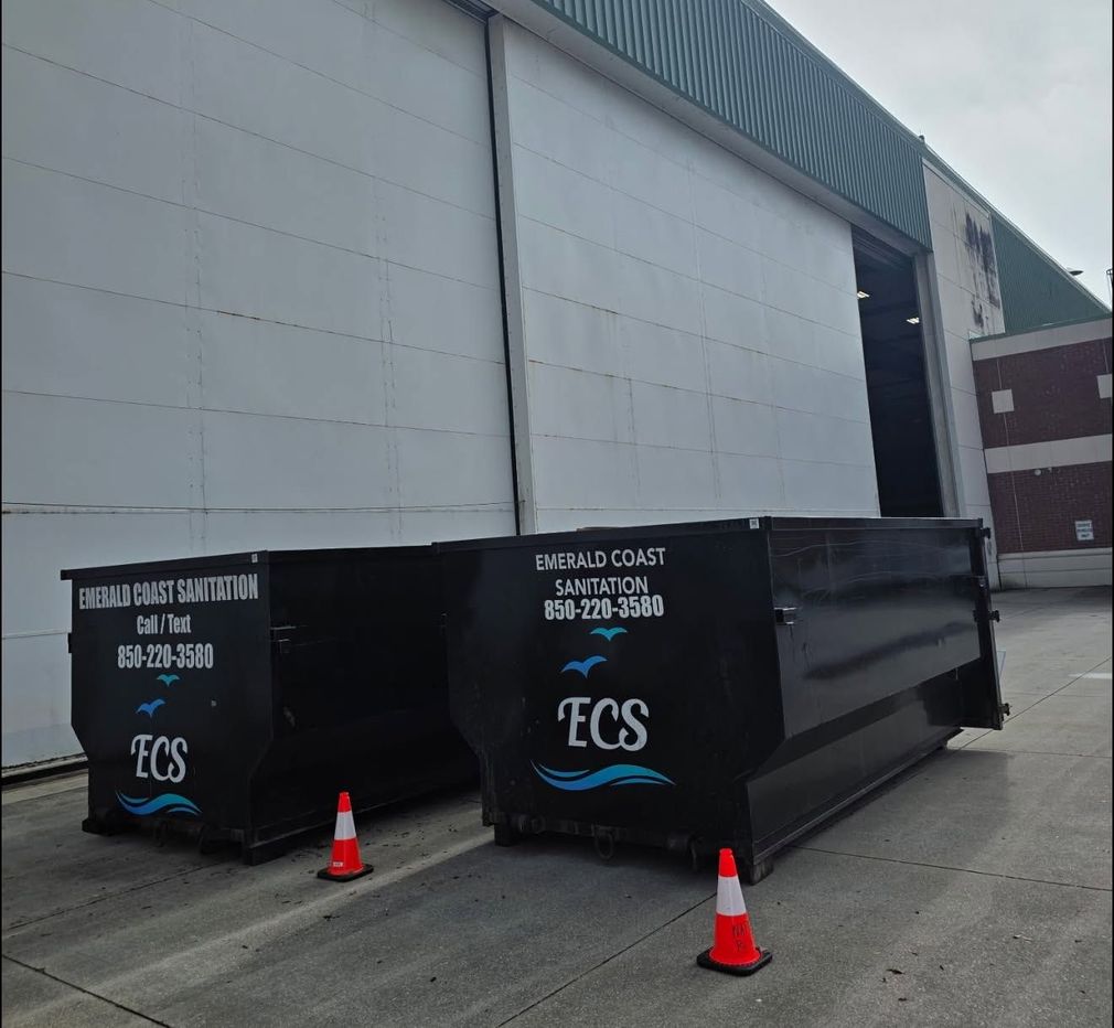Two black ECS dumpster containers by a warehouse loading bay, with orange cones in front.